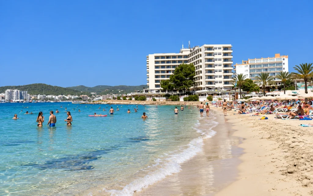 Es Pouet Beach in San Antonio, Ibiza, with tourists swimming in crystal-clear turquoise waters, sunbathers on the sandy shore and beachfront hotels in the background.